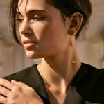 Woman in elegant black outfit wearing gold earrings and necklace from Beeline's fashion jewelry collection, showcasing the style of accessories distributed to 9,000+ retail locations