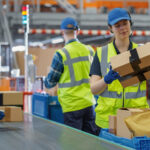 Warehouse employees in safety vests sorting and assembling packages on a modern fulfillment center conveyor line, demonstrating professional kitting operations