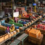 Fulfillment center employees organizing and packing colorful boxes of artisan products in a temperature-controlled warehouse, representing the custom order processing Evans Distribution Systems provides for Bon Bon Bon's chocolate fulfillment operations